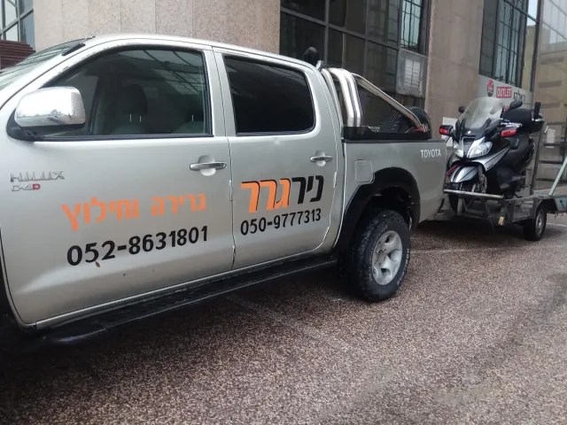 טנדר ניר גרר עם עגלה ועליה קטנוע, ליד בניין